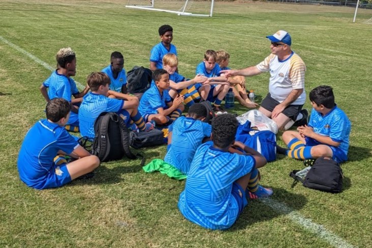 Young people from a refugee background sit on a sports field with other players and a coach